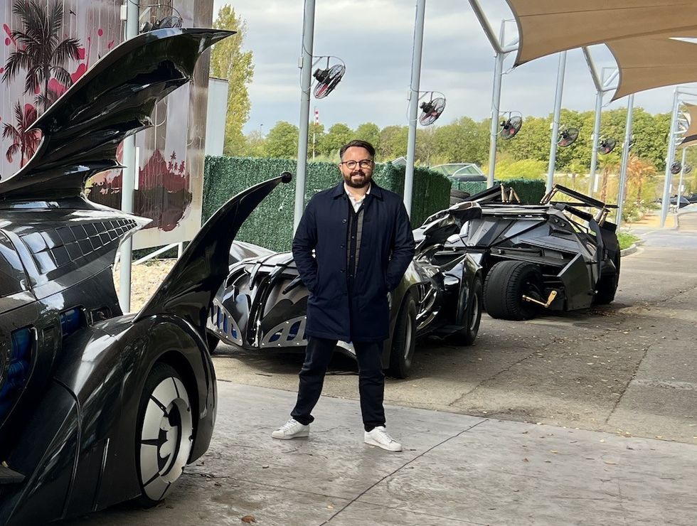 Man in a coat stands among futuristic, black, bat-themed cars.