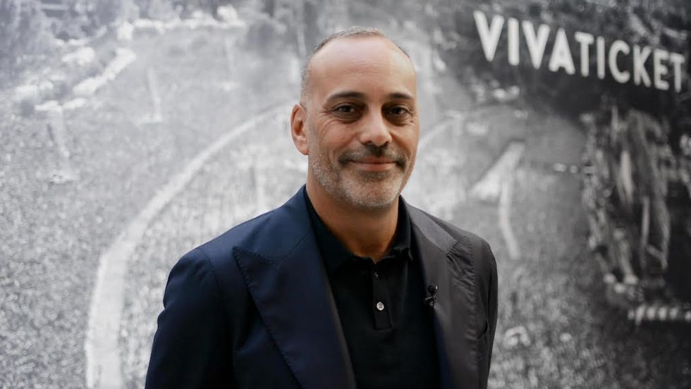 Man in a dark suit standing in front of a wall with "VIVATICKET" text.