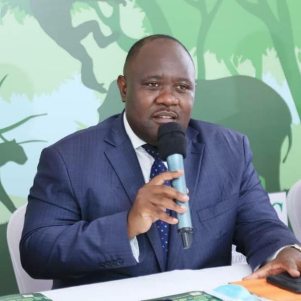 Man in a suit speaking into a microphone at an event with a nature-themed backdrop.
