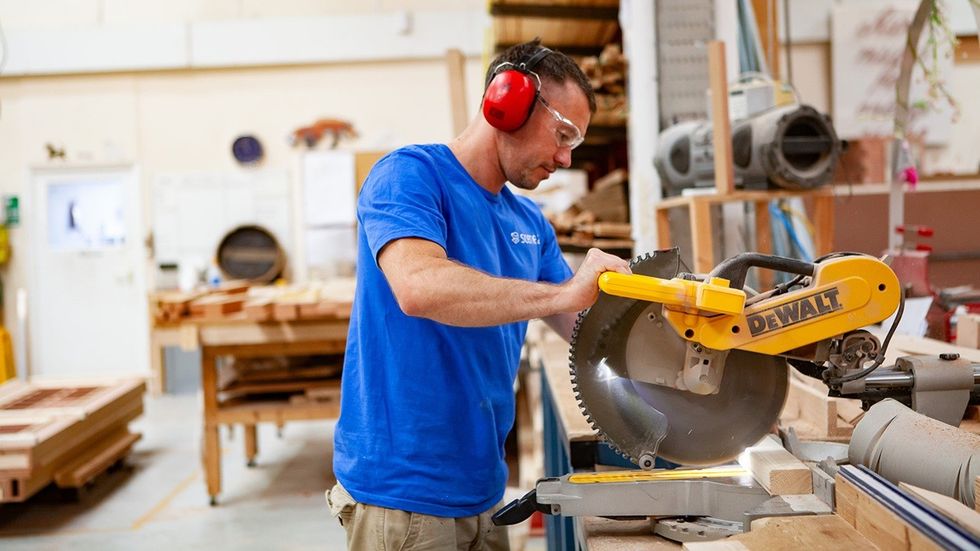 Man in a workshop using a miter saw, wearing safety gear.