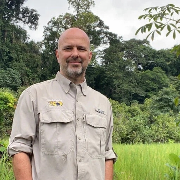 Man in beige safari shirt standing in lush, green jungle setting.