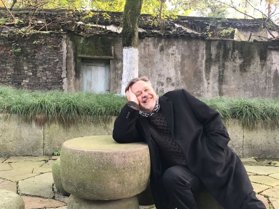 Man in black coat smiling, reclining on stone seat in front of rustic wall and greenery.