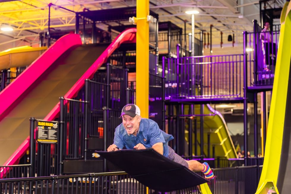 Man in blue shirt enjoying a slide at an indoor playground.