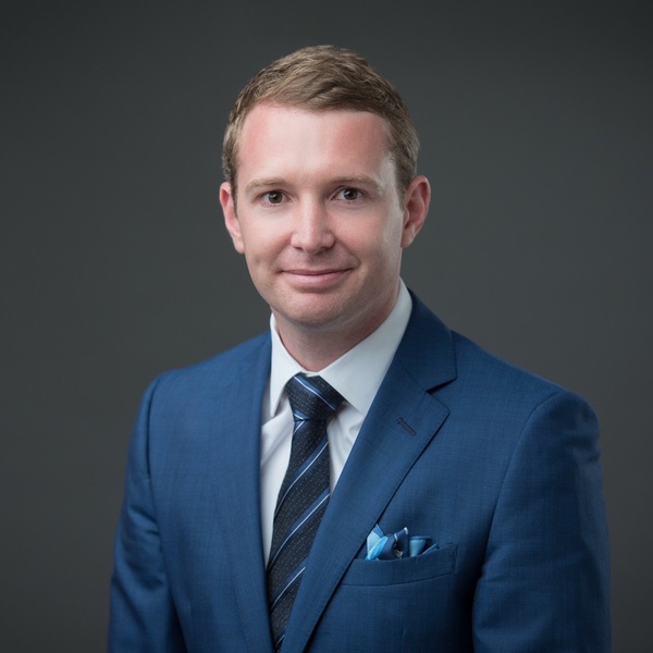 Man in blue suit and tie smiling with a neutral gray background.