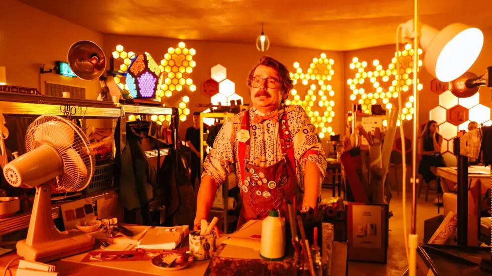 Man in colorful room with hexagonal light patterns and varied decor.