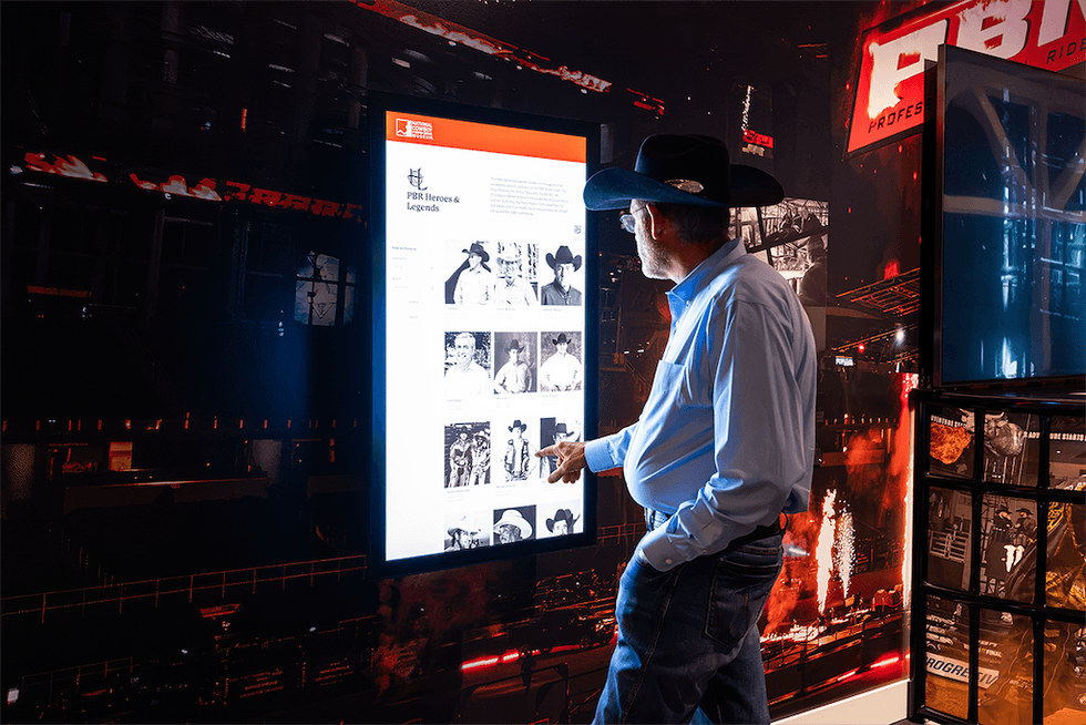 Man in cowboy hat interacts with "PBR Heroes & Legends" touchscreen display.