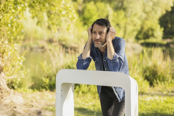 Man in denim jacket leaning on white Losonnante device in a lush, green park setting.