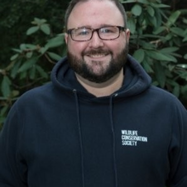 Man in glasses and hoodie with "Wildlife Conservation Society" text, outdoors with foliage.