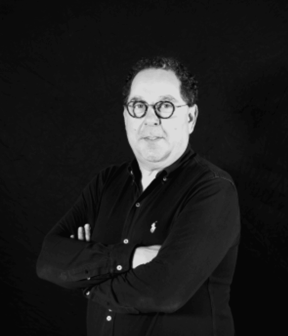 Man in glasses with arms crossed, wearing black shirt, against dark background.