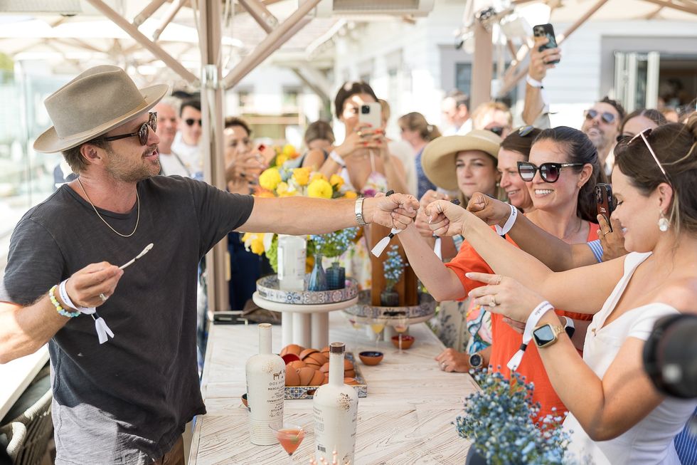 Man in hat fist-bumps fans at outdoor event, surrounded by drinks and taking photos.