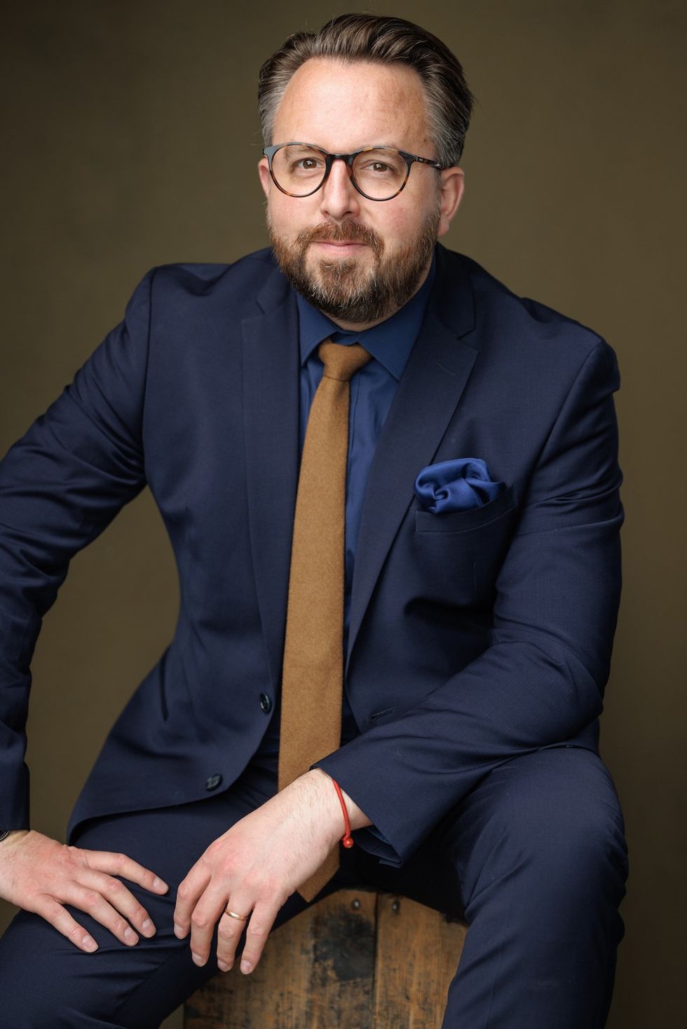 Man in navy suit and glasses with brown tie, sitting against a dark background.