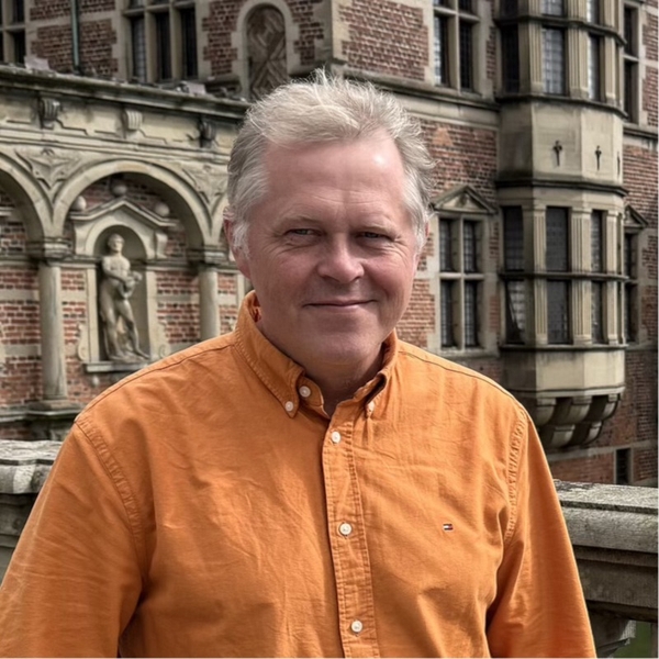 Man in orange shirt stands before ornate brick building.
