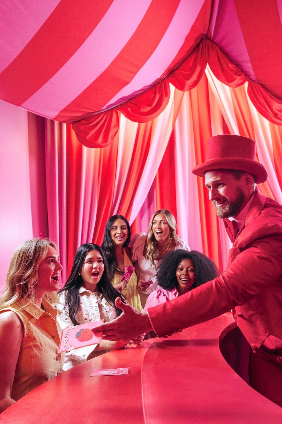 Man in red suit greets smiling women under a pink and red circus tent.