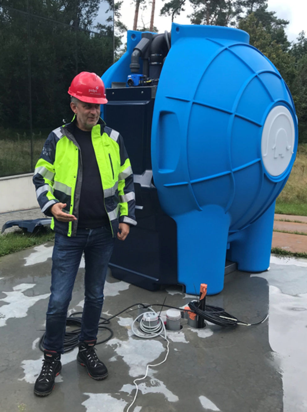 Man in safety gear stands near large blue industrial equipment outdoors.