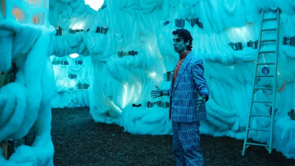 Man in striped suit in a surreal, glowing blue room with book shelves and a ladder.