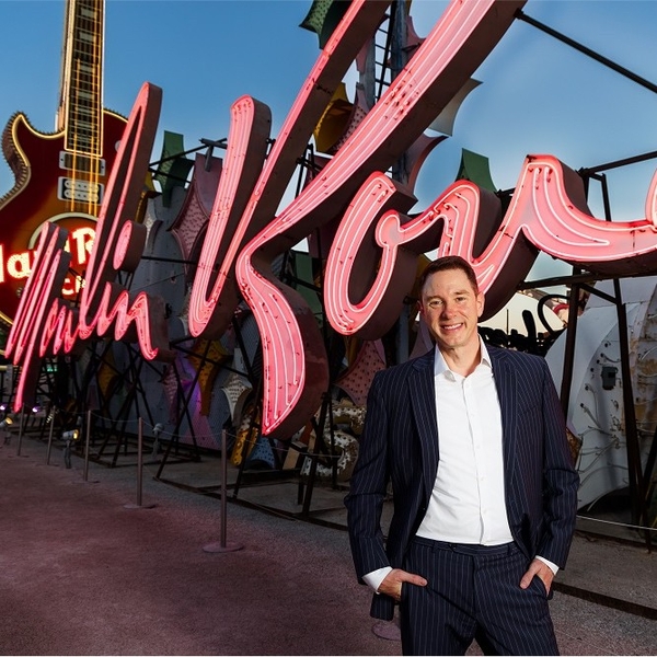 Man in suit smiles in front of large pink neon sign and guitar backdrop.