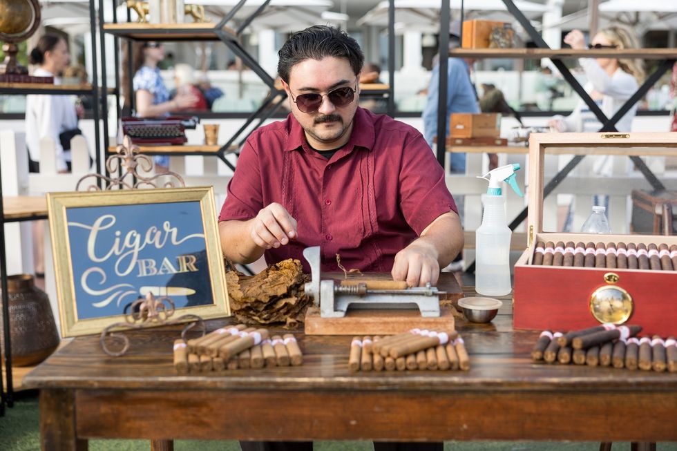 Man in sunglasses hand-rolling cigars at a cigar bar table.