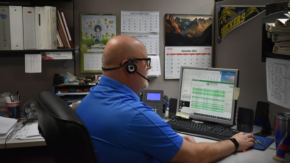 Man named T Hay in blue Chance Rides shirt and headset working on computer in an office cubicle.