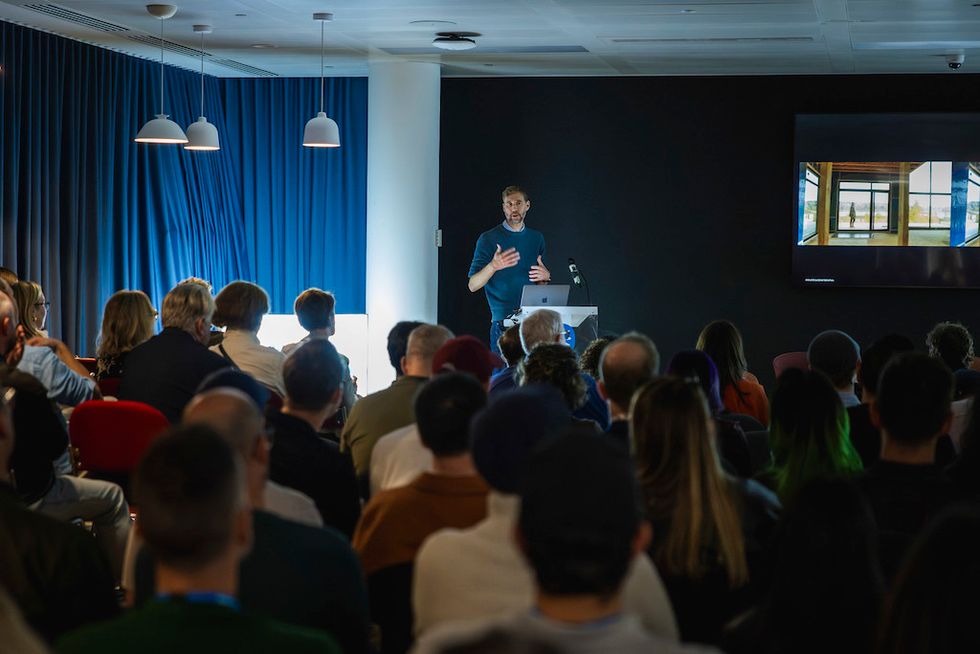 Man presenting to a seated audience in a dimly lit room with a screen showing an image.