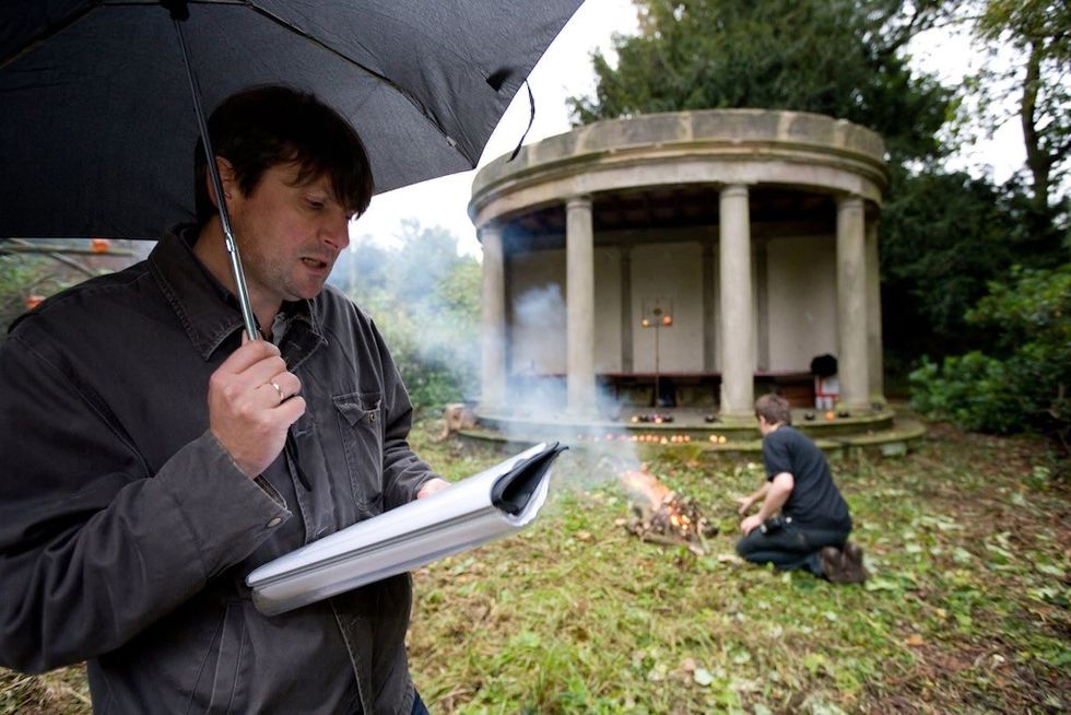 Man reads under umbrella by a fire; temple-like structure in the background.