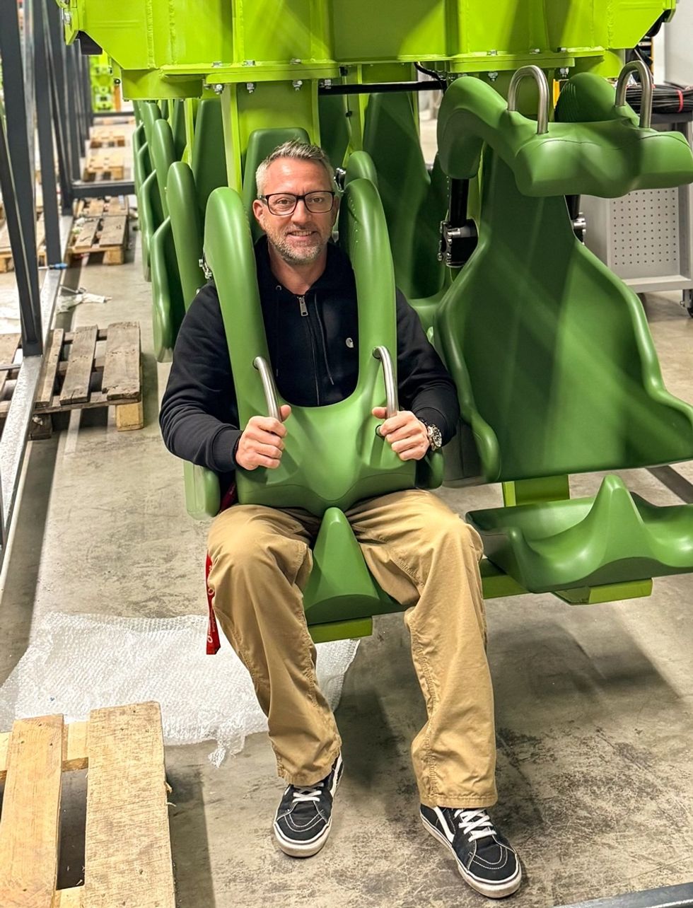 Man sitting in a green roller coaster seat, smiling indoors.