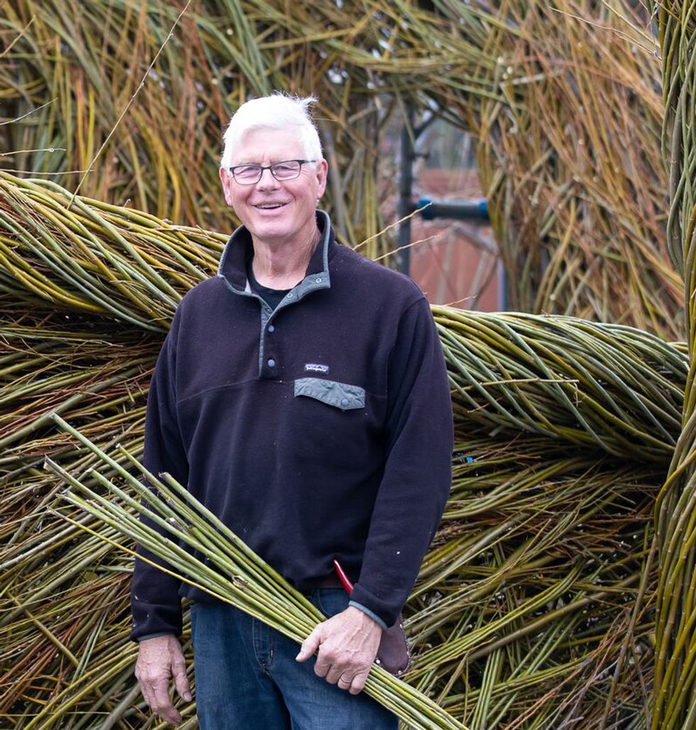 Man smiling, holding willow branches, standing by a large willow structure.