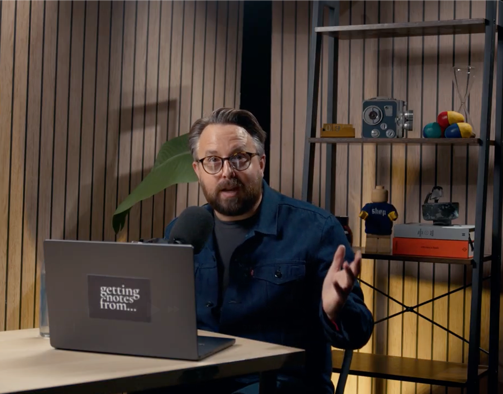 Man speaking at a desk with a laptop and microphone, shelf with decor in the background.