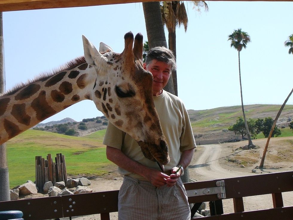 Man standing next to a giraffe at a safari park, with trees and hills in the background.