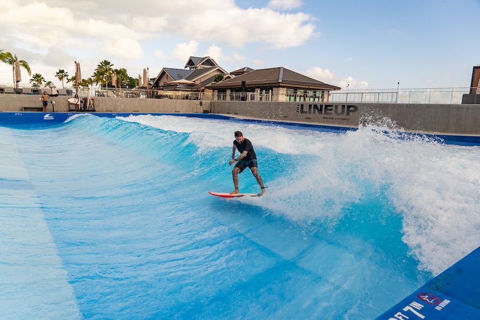 Man surfing on an artificial wave at a wave pool, with palm trees in the background.