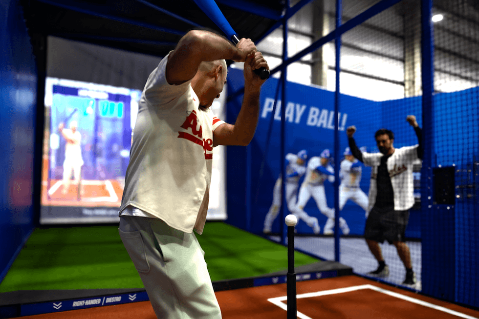 Man swinging bat at indoor batting cage, while another cheers enthusiastically nearby.