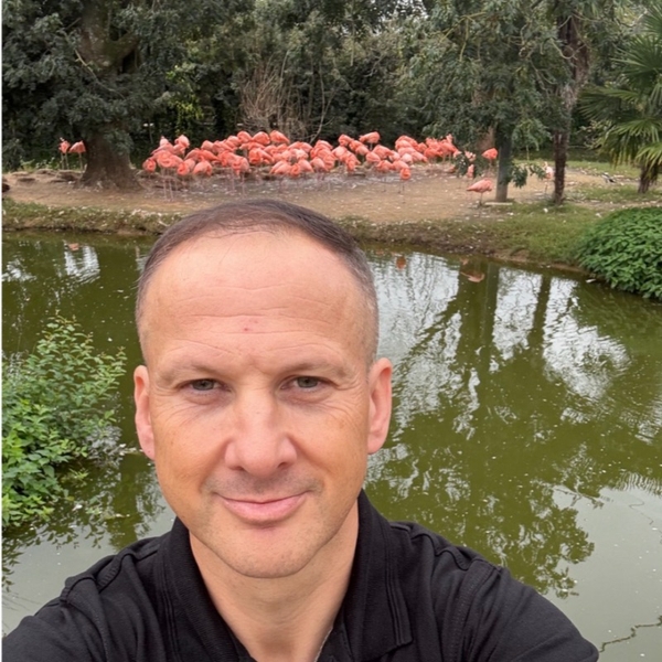 Man taking a selfie with a flock of flamingos by a pond.
