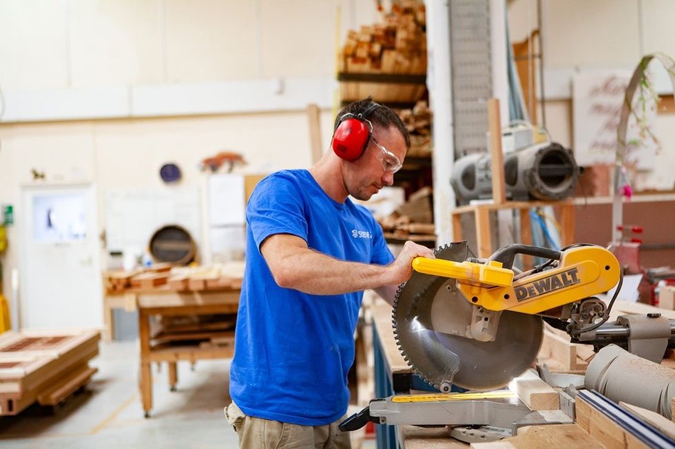 Man using a miter saw in a woodworking shop, wearing safety gear.