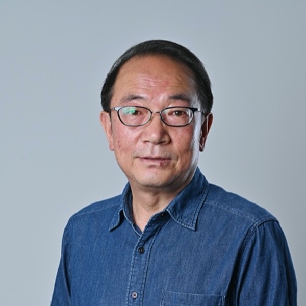 Man wearing glasses and a denim shirt, standing against a plain background.
