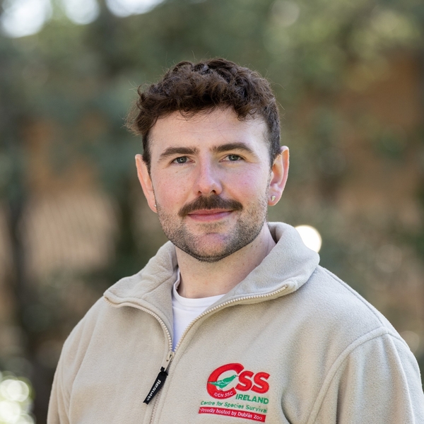 Man with a mustache in a beige jacket smiling outdoors.