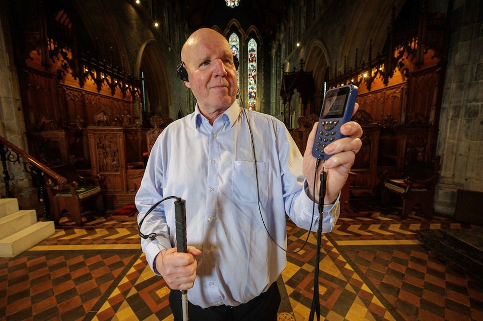 Man with assistive devices standing in a church, wearing headphones, holding a cane and gadget.