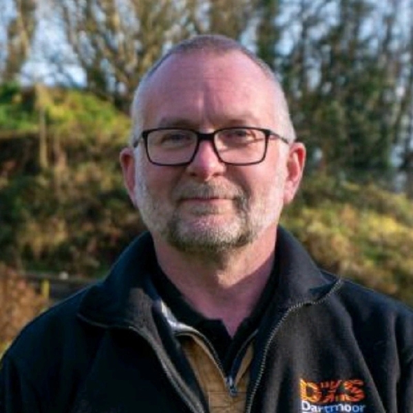 Man with glasses and beard outdoors, wearing a dark jacket with a logo.