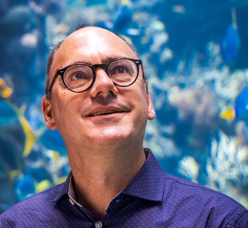 Man with glasses in front of an aquarium backdrop, wearing a blue polka dot shirt.