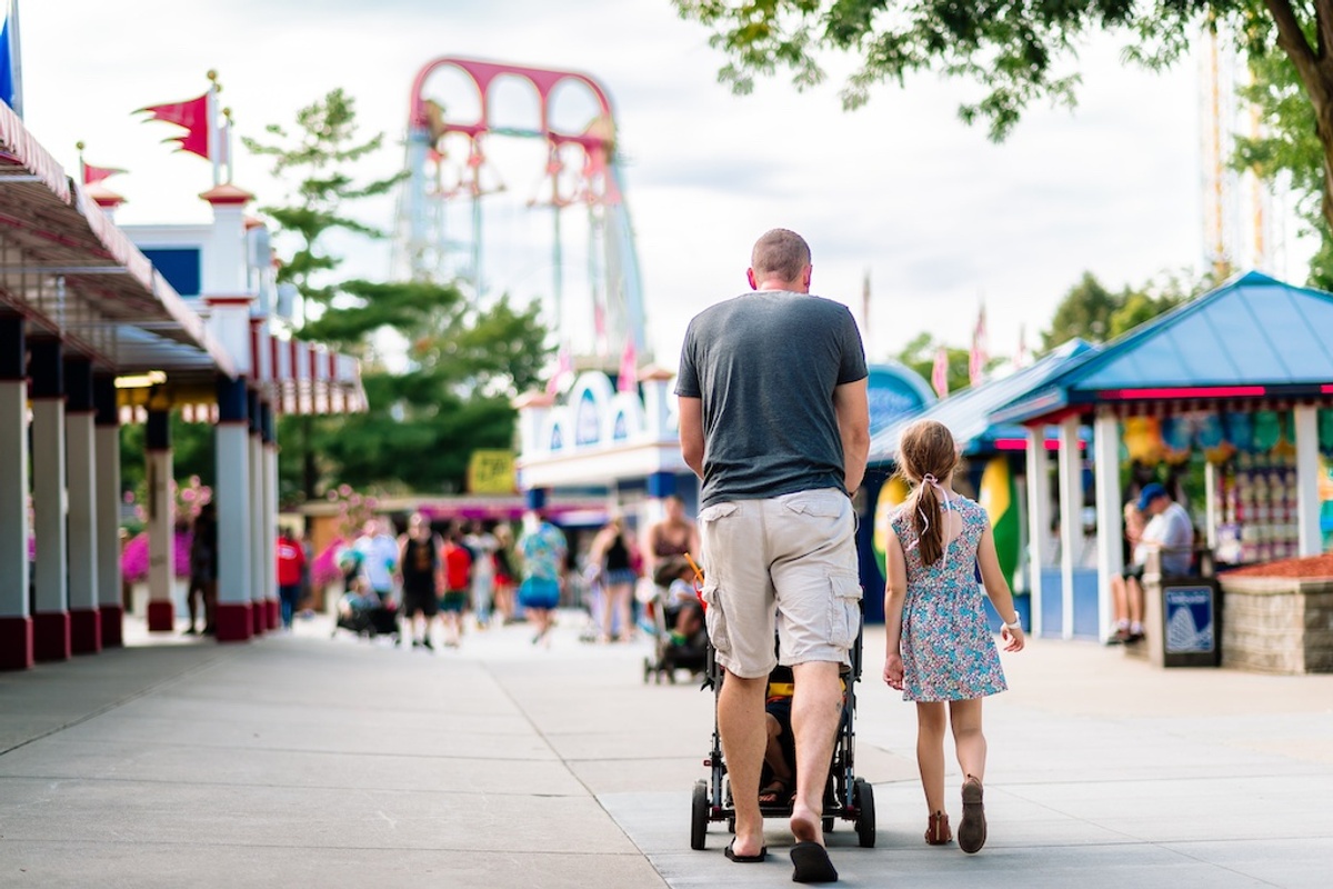 Man with stroller and child walking in amusement park, roller coaster in background.