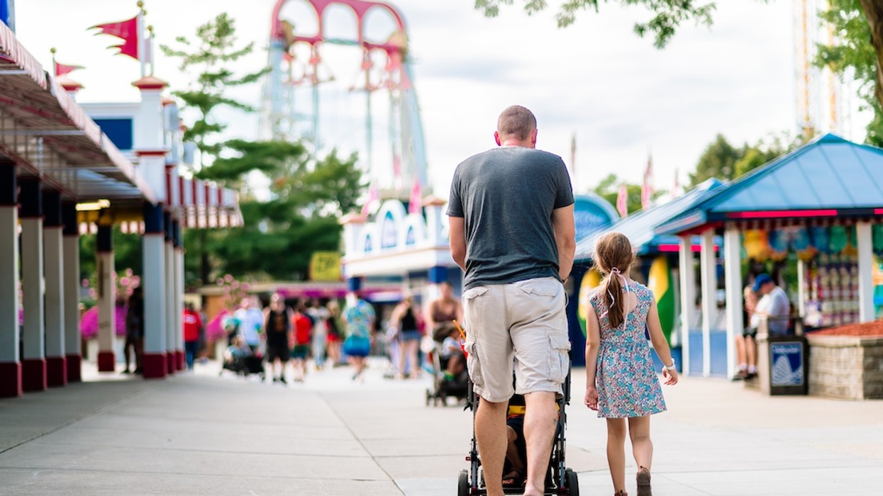 Man with stroller and child walking in amusement park, roller coaster in background.