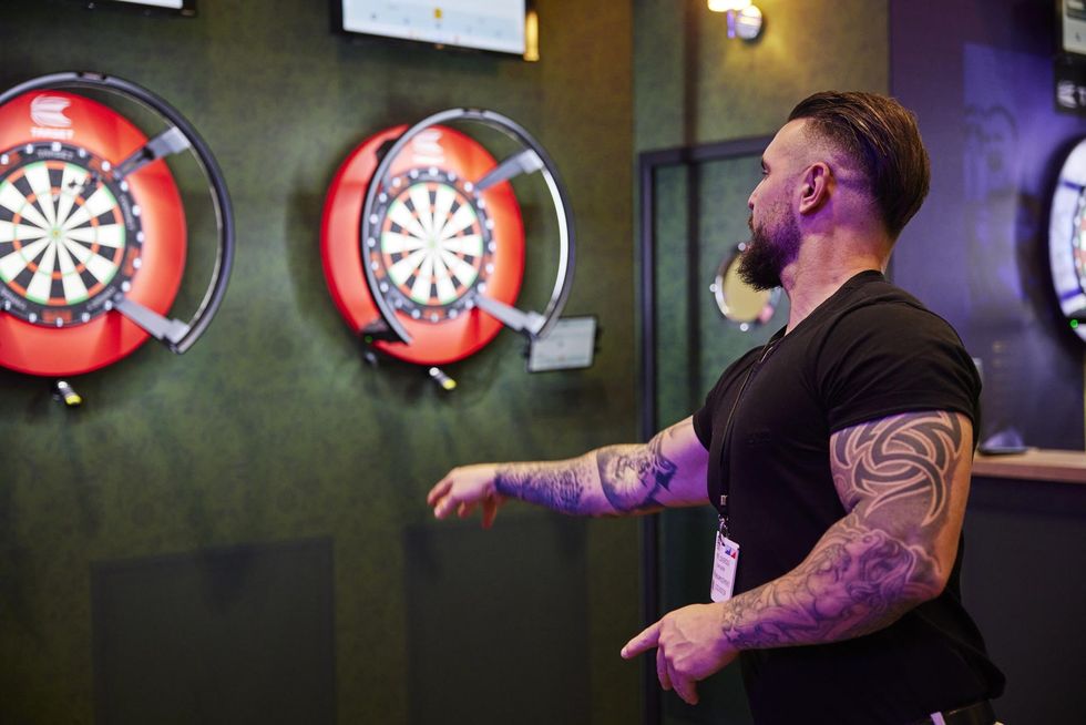 Man with tattoos playing darts indoors, aiming at two electronic dartboards.