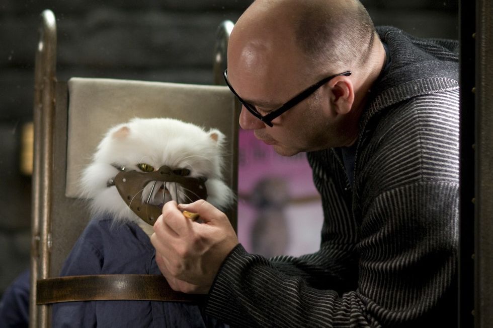 Man working on a cat puppet wearing a muzzle, seated on a chair.
