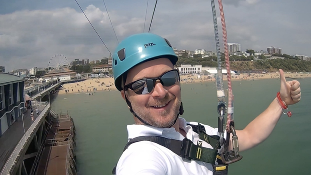 Man zip-lining over water, wearing helmet and sunglasses, beach and buildings in background.