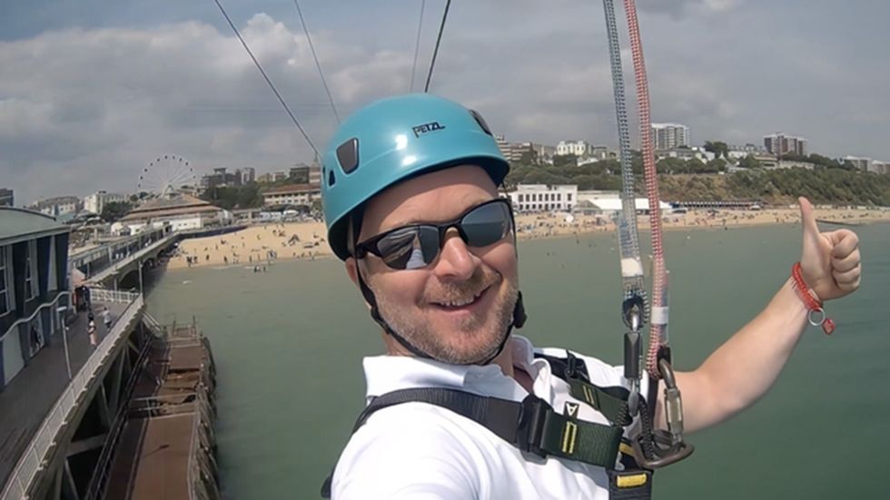 Man zip-lining over water, wearing helmet and sunglasses, beach and buildings in background.