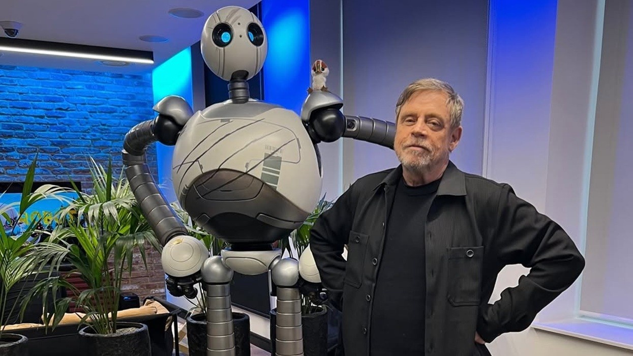 Mark Hamill posing with Roz from The Wild Robot in a room with blue lighting and plants.