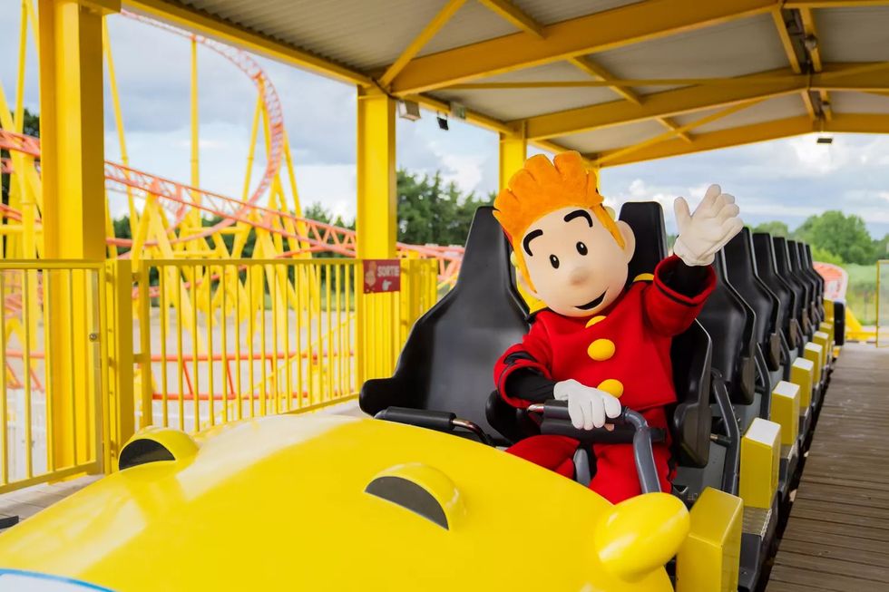 Mascot in red suit waves from yellow roller coaster car at amusement park called Parc Spirou