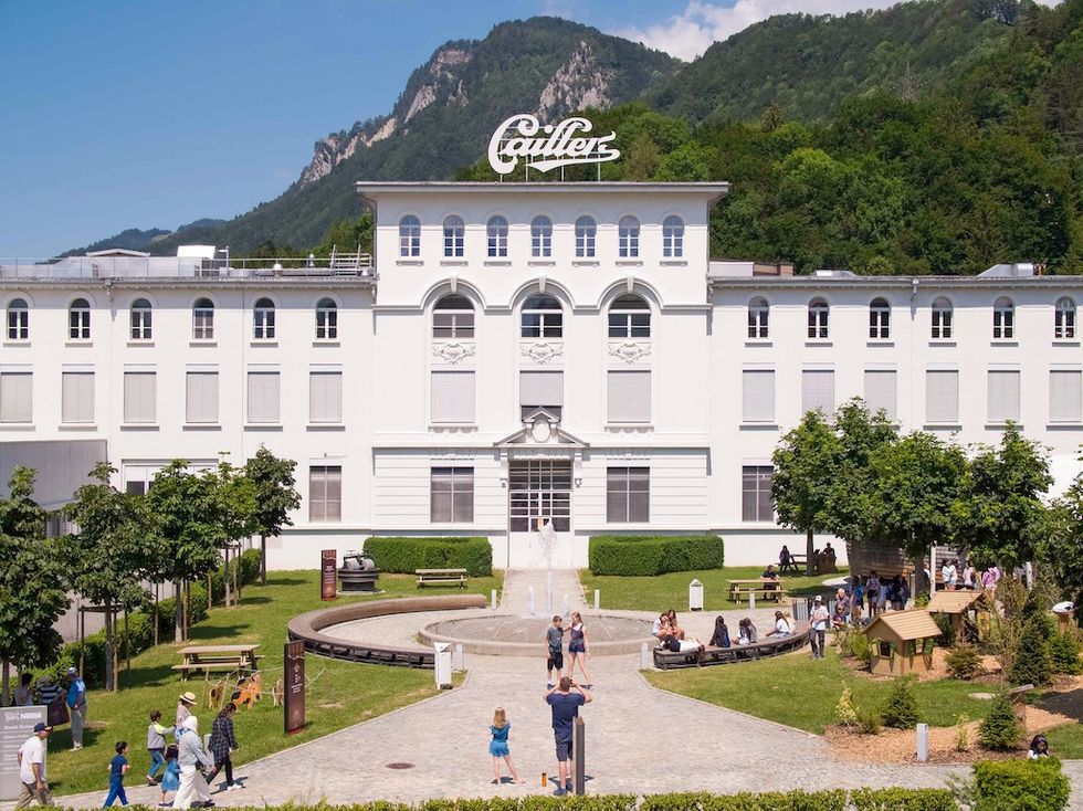 Masion Cailler - white building with "Cailler" sign, surrounded by trees and people, with mountains in the background.