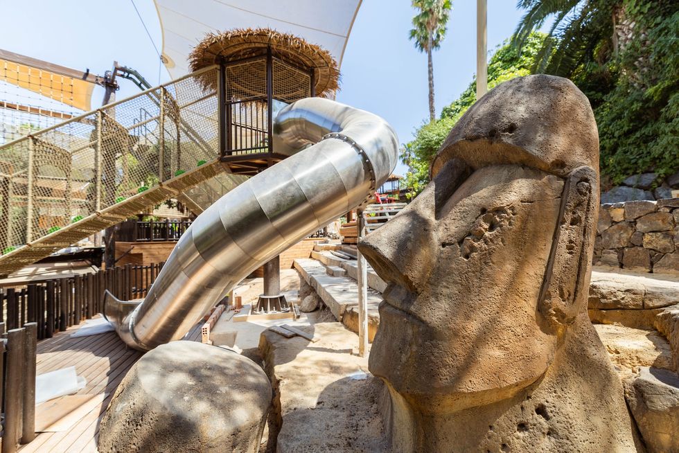 Metal slide beside a large stone sculpture, surrounded by tropical trees at PortAventura World