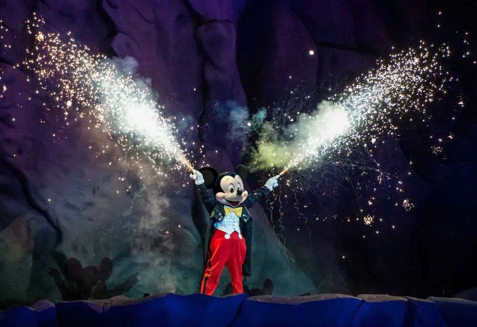 Mickey Mouse with sparklers on a stage, fireworks illuminating a rocky background.