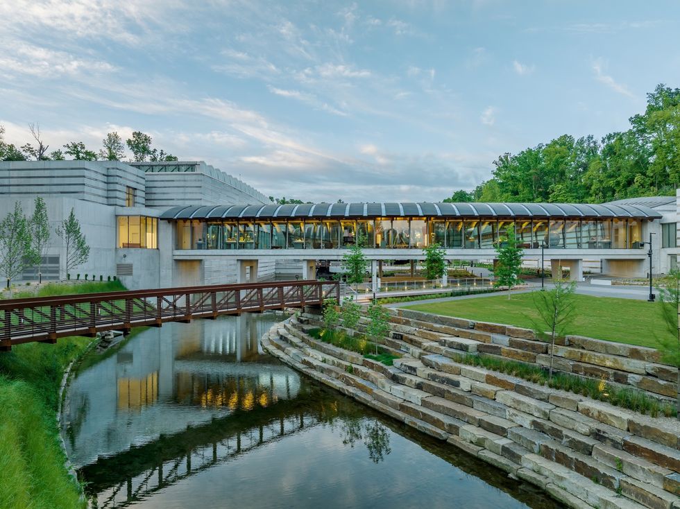 Modern building called the Crystal Bridges Museum of American Art by a stream with a wooden bridge, surrounded by trees and tiered landscaping.