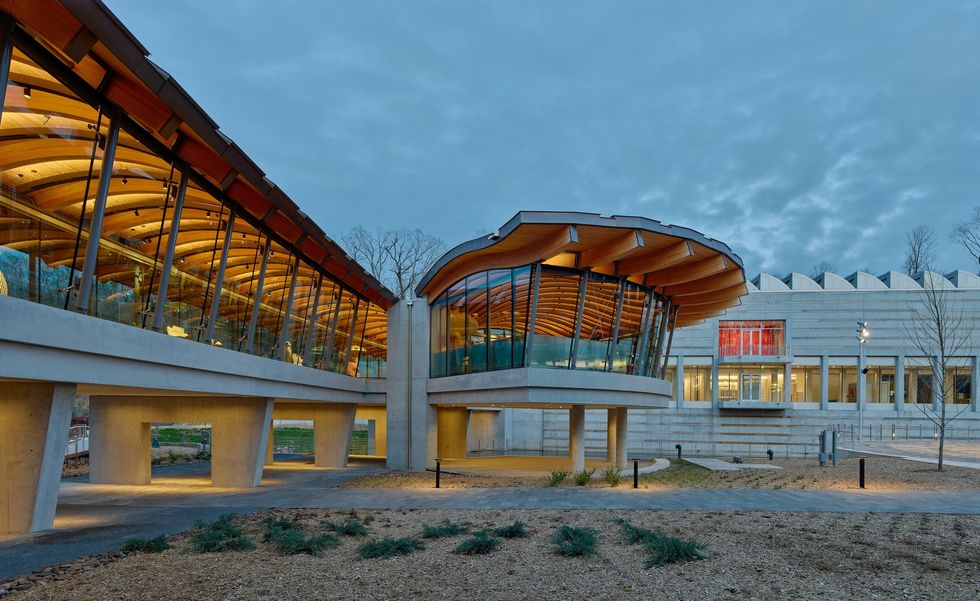 Modern building called the Crystal Bridges Museum of American Art with curved glass and wooden roof, illuminated at dusk.